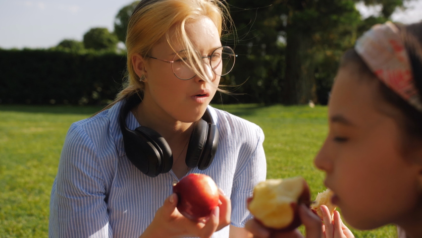 Schoolchildren of different ages are eating red apples on a green lawn. Close-up of a girl biting a red apple.