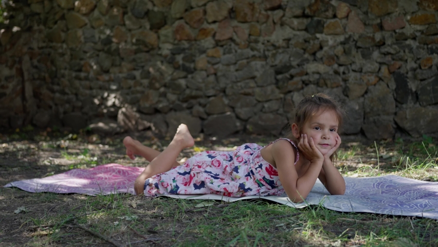 A little beautiful girl plays on a yoga mat, in the summer in the garden, she has fun and rejoices, repeats gymnastic exercises, smiles. slow motion