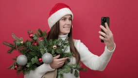 Portrait of good-looking young brunette female in Santa Claus hat holding bouquet of spruce branches do selfie shot on mobile cell phone post photo on social network, posing over red studio background