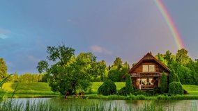 Wooden hut on lake banks in bucolic and idyllic landscape with colorful rainbow in background. Time-lapse.