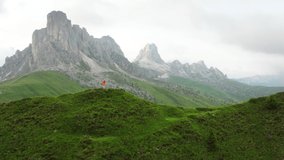 4K drone flying around person wearing orange windbreaker catching majestic Italian Alps afar in background. Hiking, traveling, active lifestyle, exploring nature concept.