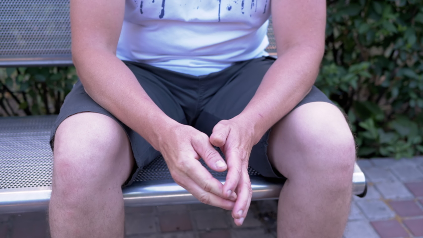 Male Rubbing Hands Sitting on a Bench in the Urban Park. A moment of waiting, excitement, anxiety, and emotional stress, trouble, nerves, and dread. Massage of hands, joints, fingers, skin. Outdoors.