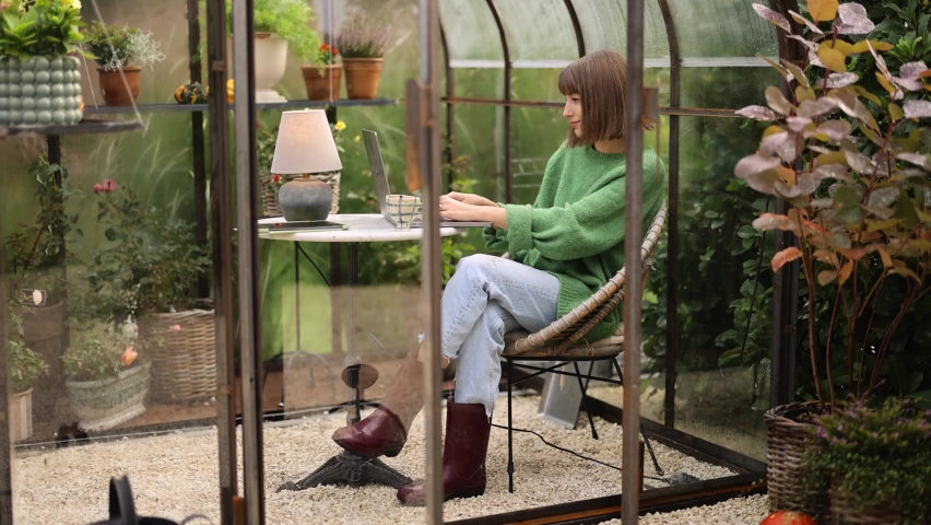 Young woman works on laptop while sitting by the round table in glasshouse with plants and flowers at backyard. Work from home at cozy atmosphere on nature