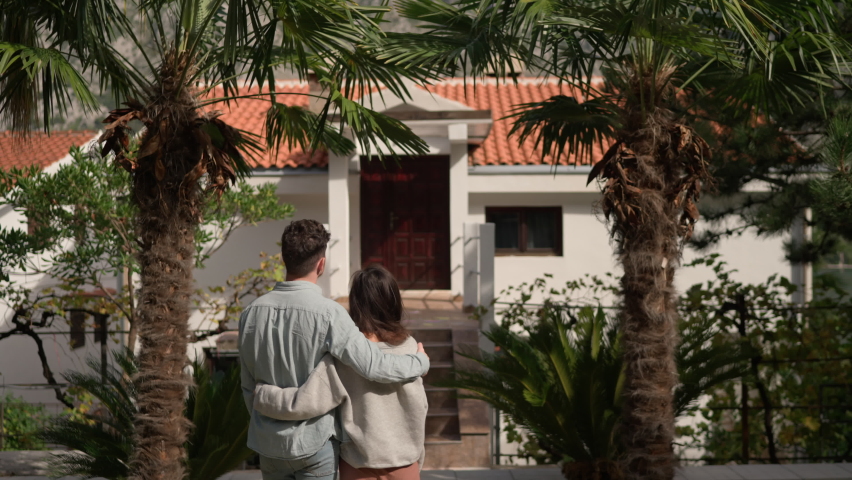 Rear on Asian American young just-married couple standing at yard of house, hugging and observing second floor. Back view on man and woman in hugs moving in new home.