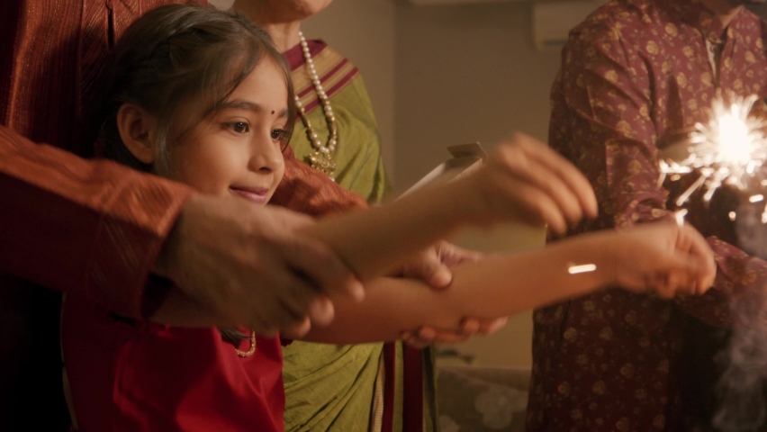 Ethnic Indian Hindu happy smiling cute young girl or female kid in traditional clothes or attire playing with firecrackers or sparklers with the help of family elders or parents during Diwali festival | Shutterstock HD Video #1095190859