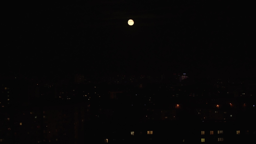 Large round yellow moon moves over the big city at night, time lapse video. Clouds are moving in front of Earth satellite. Lights are lit in some windows of high-rise residential buildings.