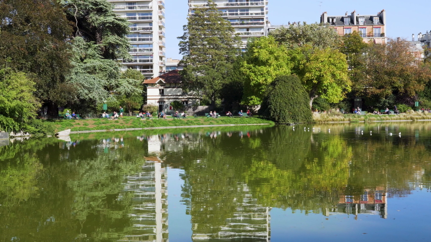 Pan over the lake of Parc Montsouris in Paris with Birds, reflection of buildings and Parisians walking and sunbathing. Shot on a sunny day at early autumn - Paris, France