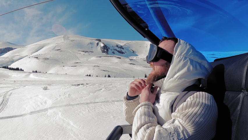 Bearded mid adult man skier in pretextive goggles and helmet sitting in ski lift at resort in Alps