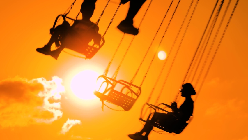 Unrecognizable silhouettes of people ride on the chained carousel against warm bright orange sky at sunset with sun lens flares at amusement park. Summer, fun, entertainment and holiday concept | Shutterstock HD Video #1095216163