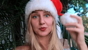 Funny young beautiful smiling girl in Santa's hat celebrates Christmas on vacation, looking at the camera on the background of plants. Woman grimacing, playing with pom-pom on hat and fooling around