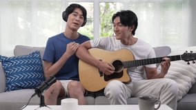 Young Asian gay couple blogger vlogger and online influencer recording musical video content playing guitar and singing at home. LGBT gay couple performing and shooting clip for social media.