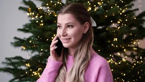 A beautiful girl in a pink sweater stands against the background of a Christmas tree in a cozy home interior and talks on the phone. Slow motion horizontal video