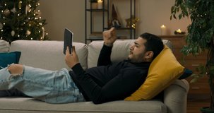 A lonely guy with dark hair lies on sofa on yellow pillow and holds tablet in hand. He is wearing black sweater and ripped jeans. The boy emotionally watches boxing, comments and gestures with hands.
