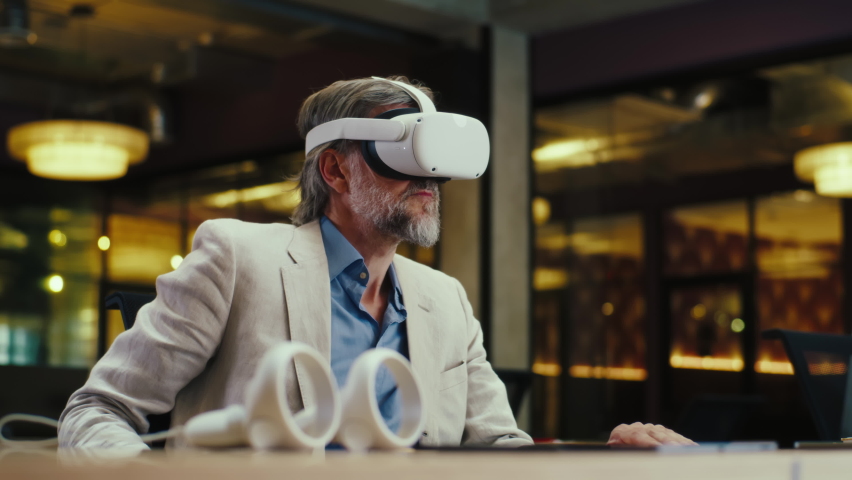 Mature man sitting at workplace, wearing VR headset, watching data in virtual reality, testing device. Work in modern office of hi-tech company. Cyberspace digital technology - Powered by Shutterstock - Get 15% off with code: PIKWIZARD15