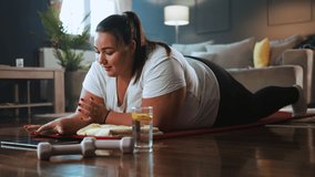 Young overweight woman doing a plank exercise having an online class with a personal trainer on a video call from home