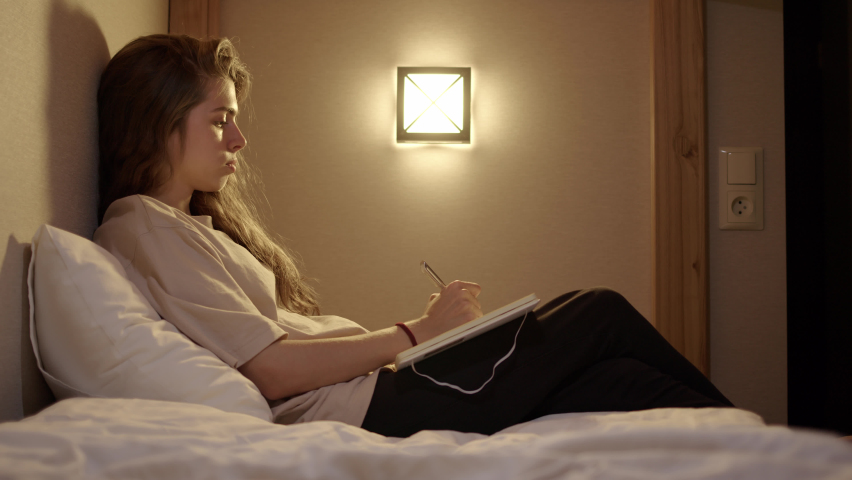 Attractive young woman in white t-shirt writes entries in diary or notebook while lying on bed with lamp on wall and moonlight. girl makes notes in book and studies. evening writing letter to friend.