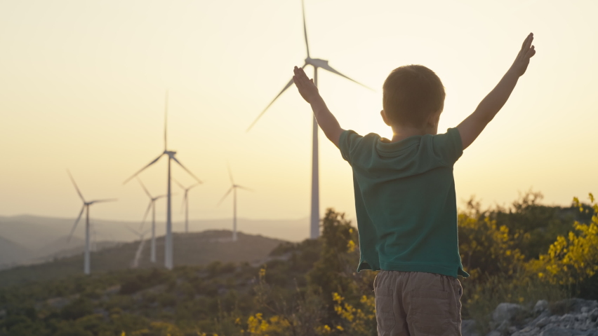 The boy rejoices and waves his hands at the sunset near the windmills. A child and a source of renewable ecological wind energy in the mountains. Production of electricity in a wind farm. 