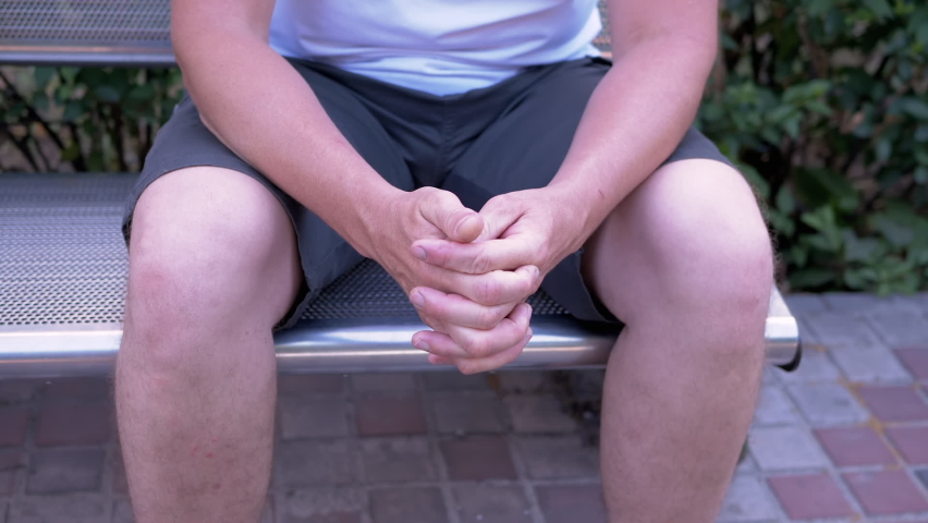 Male Rubbing Hands Sitting on a Bench in the Urban Park. A moment of waiting, excitement, anxiety, and emotional stress, trouble, nerves, and dread. Massage of hands, joints, fingers, skin. Outdoors.