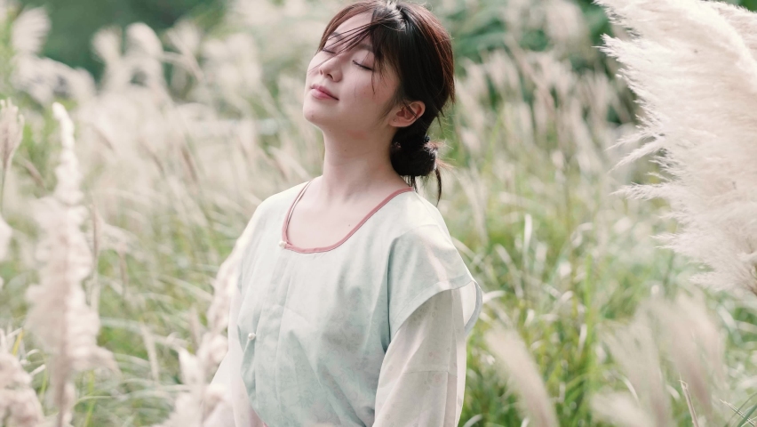 Beautiful Chinese girl in traditional clothes enjoy the breeze in reeds field in autumn day, eyes closed then smiling and looking at camera, 4k slow motion footage.