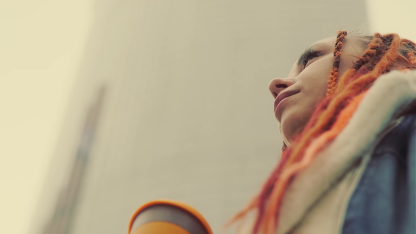 From below of woman with dreadlocks in warm clothes drinking coffee from cup, looking up, standing on street.