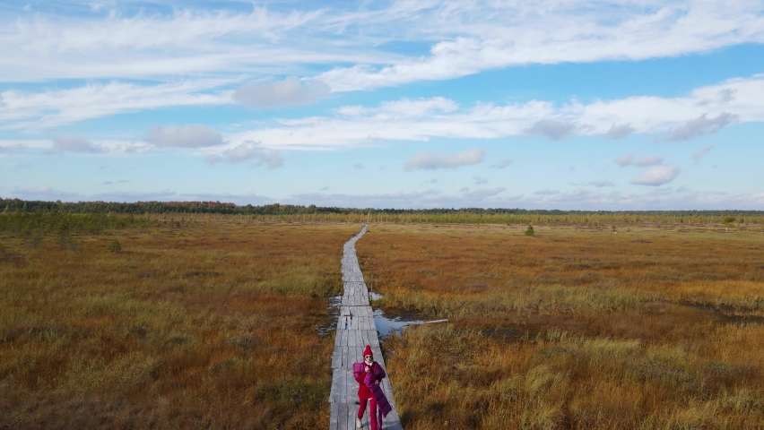 fashionable woman in a red sweater , hat and scarf walks along a trail of planks through a swamp in autumn in Yelnya in Belarus