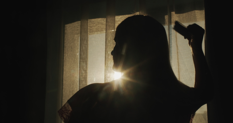 Silhouette of a middle-aged woman combing her hair at the window