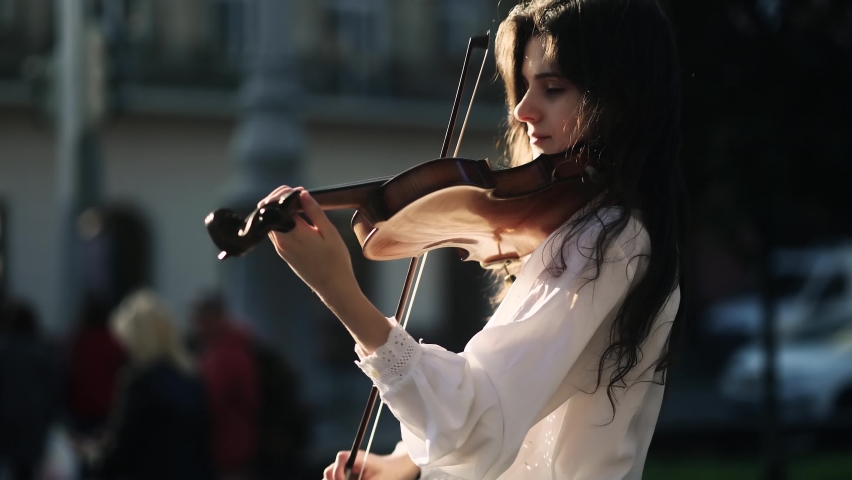 Young street musician playing classical wooden violin on the urban city street. Talented violinist with incredible sunset on the background.