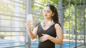Portrait of charming indian girl in black sport wear making a video call smiling, talking, waving her hand  after workout near the stadium. Smiling young woman have a call. 