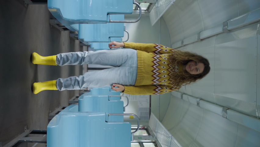 Curly-haired tourist in yellow boots stands between blue seats in empty train. Young woman looks in camera smiling on way to forest vertical shot