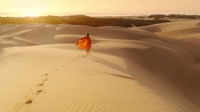 Young beautiful girl in orange linen safari dress walking along sandy dunes in scenic vast desert at ocean coast. 4K Slow Motion aerial back view of traveler hiking barefoot by desert at golden sunset - Powered by Shutterstock - Get 15% off with code: PIKWIZARD15