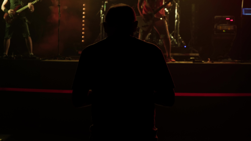 unrecognizable Sound Engineer adjusts sound for rock concert in large event hall. Rear view silhouette of man in spotlights with artists and guitarists on stage in background.