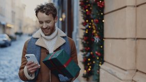 Caucasian happy man going home from the christmas gift shop holding xmas gift using smartphone, texting with friends or relatives. People and technology concept - Powered by Shutterstock - Get 15% off with code: PIKWIZARD15