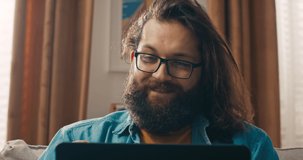 Close-up shot of face of bearded hipster man with glasses browsing internet on laptop, online stores in search of gifts for loved ones, home accessories.