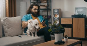 A bearded man browses the web on a smartphone in the company of dog. A contented young boy uses phone while sitting on the living room couch smiling while stroking pet.