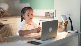 Happy schoolgirl at table online lesson. Girl child does homework. Schoolgirl at desk performs task of teacher. Online lesson with laptop. Student in school classroom. Home Internet for teaching child