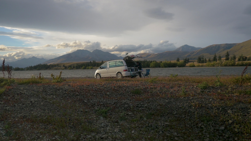A woman cooks next to her van in front of a lake, in New Zealand