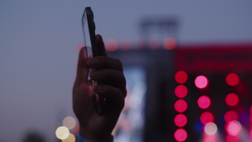 Closeup of hand holding smartphone device vertically at the music festival, using camera app to shoot video of the crowd in front of the stage, evening outdoor event, side view Royalty-Free Stock Footage #1095284387