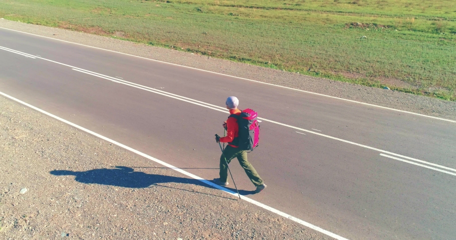 Aerial 4K UHD view. Low altitude flight over hitchhiker tourist walking on asphalt road. Huge rural valley and sunny meadow at summer day. Backpack hiking guy.