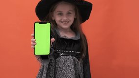 Halloween girl on orange background. Kid in a witch costume. Happy Halloween. A girl is holding a smartphone with a green screen. Internet shop. Online shopping with sales.