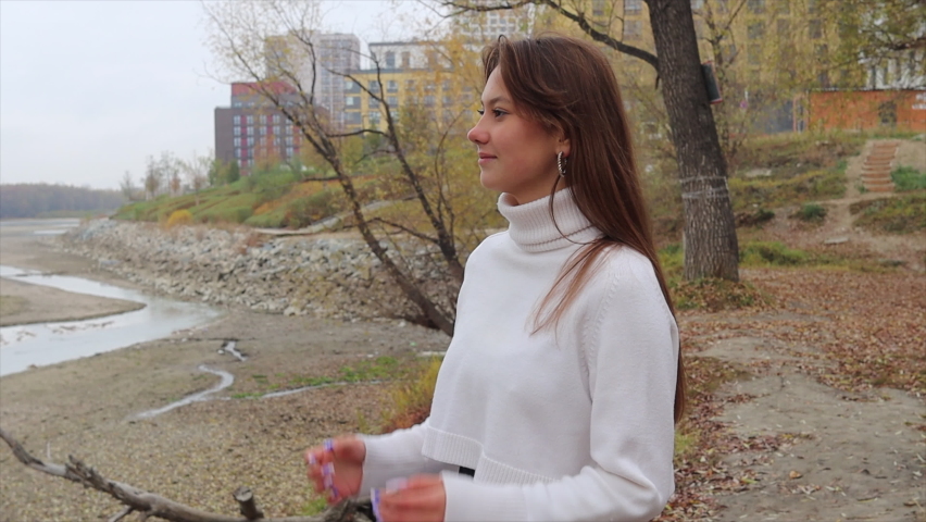 Young frozen woman straightening clothes in park. Autumn. Cloudy day.