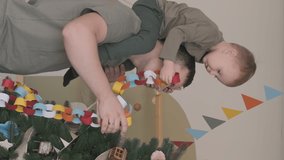 Vertical video. Young Caucasian Father with his Little Son Decorating Christmas Tree at Home, Child Sitting on the Shoulders of his Dad. Happy Childhood, Christmas and New Year Celebration Concept