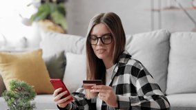 Relaxed Female shopper in glasses holding credit card and smartphone sitting on couch at home. Young woman customer using instant easy mobile payments making purchase in online store. Donation