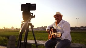 Folk musician playing guitar filming vlog on smartphone at sunset