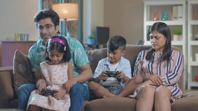 A happy smiling Indian Asian family of mother, father, son, and daughter playing a video game together sitting on a couch in an interior home. Relationship, bonding, togetherness, affection concept.