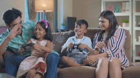 A happy smiling Indian Asian family of mother, father, son, and daughter playing a video game together sitting on a couch in an interior home. Relationship, bonding, togetherness, affection concept. - Powered by Shutterstock - Get 15% off with code: PIKWIZARD15