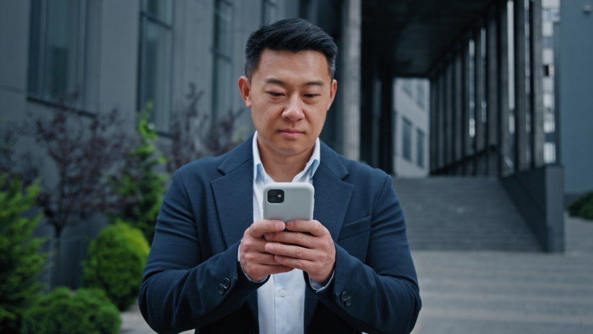 Asian mature businessman holding smartphone standing outdoors in city with smile. Middle-aged manager man CEO business leader using cell phone mobile app for working distant looking at camera smiling Royalty-Free Stock Footage #1095330189