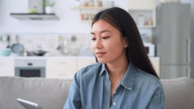 Young casual asian woman entrepreneur using digital tablet relaxing at home sitting on sofa couch. Millennial girl watching funny video in social network or chatting with friends online - Powered by Shutterstock - Get 15% off with code: PIKWIZARD15