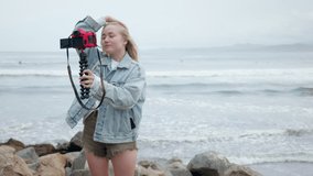 Shot of a young blonde woman standing and filming herself by camera on the ocean beach. Big Sur California coast. Waves crashing. Travel blogger on vacation. High quality 4k slow motion footage.