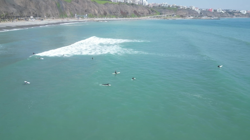 Aerial spinning birdseye view off the coast of miraflores lima in peru with surfers with surfboards in the turquoise sea in the waves overlooking the highway along the rocks on a sunny day