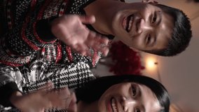 Vertical Screen: cheerful asian couple waving hi to webcam and touching each otherâ€™s face and fat belly while giving Christmas greetings in delightful video chat on sofa at home